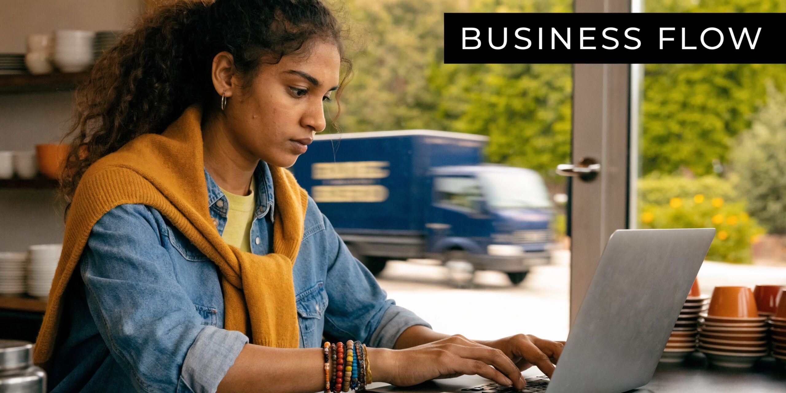 A young woman working on her laptop at a cafe with a delivery truck visible outside.